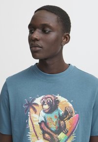 Blå bomulds t-shirt med et livligt print af en abe i solbriller, omgivet af tropiske elementer og et surfbræt-design.
