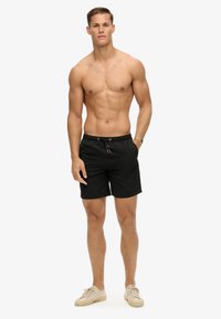 Pantaloni da nuoto neri con cintura elastica, cordoncino e tasche laterali. Il modello indossa sneakers beige. Tessuto morbido, lunghezza a metà coscia.