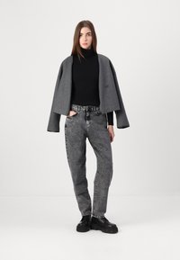 Γκρι κοντό σακάκι πάνω από μαύρο φίλτρο, συνδυασμένο με ψηλόμεσα γκρι τζιν. Το μοντέλο φοράει μαύρες πλατφόρμες, στέκεται μπροστά από ένα ουδέτερο φόντο.