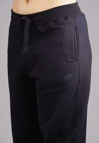Close-up of a person wearing black sweatpants with drawstring waist and side pockets, showing a small embossed logo on the thigh.