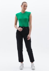 Zelený žebrovaný top bez rukávů kombinovaný s černými džíny s vysokým pasem. Bílé tenisky s černými akcenty. Minimalistické pozadí.