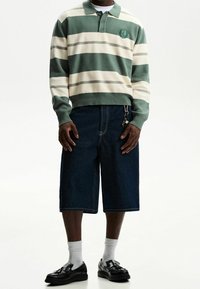 Person med grön- och vitrandig långärmad polo, mörk denimbykort med nyckelring, vita strumpor och svarta franslövar.