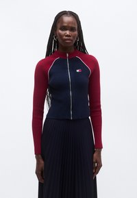 Soonikkoes lukuga kampsun mereväe sinises ja burgundia toonis, millel on poolkõrge kaelus, kontrastsed varrukate detailid ning rinnal tikitud logo.