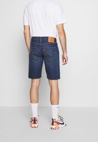 Pantalones cortos de mezclilla en azul oscuro con corte recto, dos bolsillos traseros, costuras en contraste y una etiqueta de cuero marrón en la cintura.