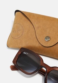 Barna Ray-Ban napszemüveg téglalap alakú kerettel, sötét lencsékkel és logóval. Egy bézs bőrtokkal kísérve, amely dombornyomott részletekkel és patentos zárással rendelkezik.