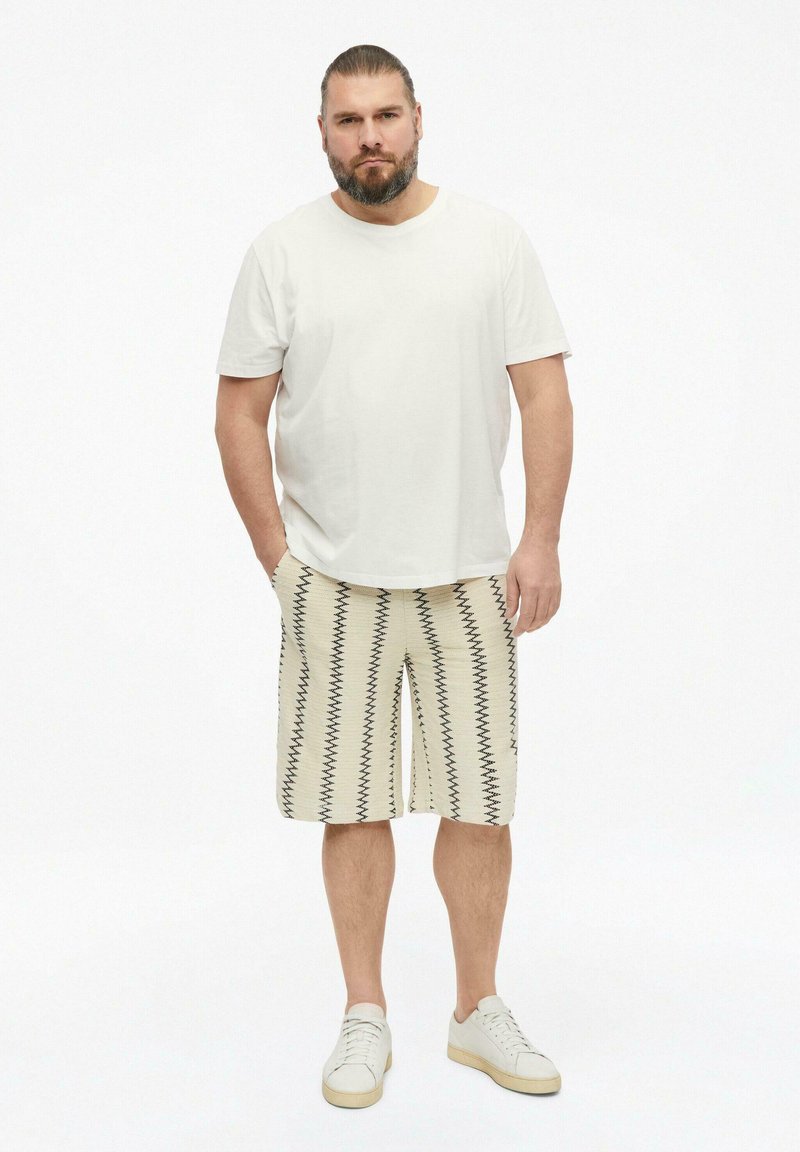 Homme avec barbe et cheveux attachés, portant un t-shirt blanc uni, un short crème à rayures noires en zigzag, et des baskets blanches, debout les mains dans les poches.