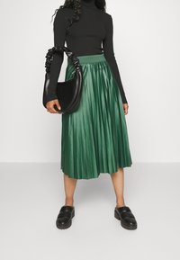 Nainen, jolla on yllään musta pitkähihainen paita, vihreä laskostettu midi-hame, mustat paksupohjaiset slip-on-kengät, ja hän pitää kädessään mustaa rypytettyä käsilaukkua.