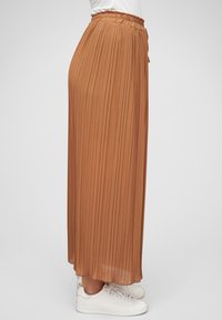 Pleated brown maxi skirt made of lightweight fabric with an elastic waistband, paired with white sneakers, shown from the side.