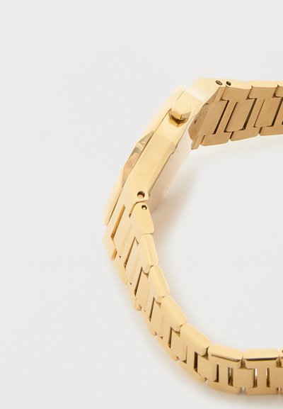 Χρυσό μεταλλικό ρολόι με δεμένη μπρασελέ και στρογγυλή κάσα, εμφανίζεται από πλευρική γωνία σε λευκό φόντο.