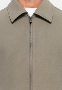Veste gris clair avec un col structuré, fermeture éclair sur toute la longueur et tissu texturé. Design simple, éclat subtil et sans motifs.