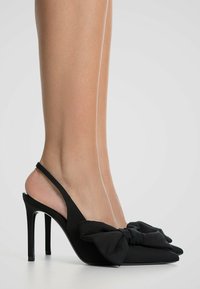 Zwarte slingback hoge hakken met een puntige neus, satijnen textuur, grote strikdetail aan de voorkant en een slanke stilettohak.