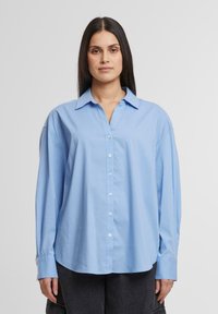 Camicia azzurro chiaro, con bottone e colletto a punta, maniche lunghe e vestibilità rilassata. Tessuto morbido e liscio con un design semplice.