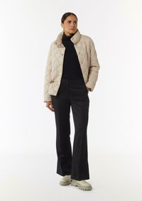 Beige gesteppte Jacke mit hohem Kragen, schwarzem Rollkragenpullover und schwarzen Schlaghosen. Modell trägt hellfarbige klobige Stiefel.