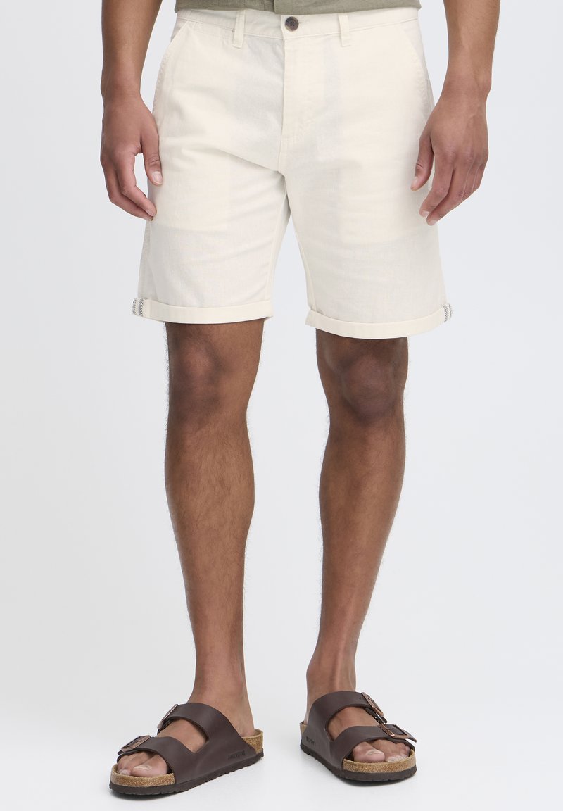 Beige knielange shorts met opgerolde cuffs, voorzien van een enkele knoopsluiting en twee zijzakken, gecombineerd met bruine sandalen.