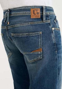 Jeans en denim bleu foncé, dotés d'une patch en cuir marron au niveau de la taille et des poches arrières avec des détails de couture distincts.