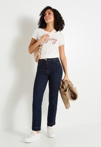 Camiseta blanca de manga corta con un diseño impreso colorido, combinada con jeans de un azul oscuro, zapatillas blancas, y sosteniendo una chaqueta y un bolso beiges.