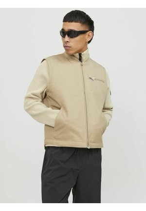 Beige vest met hoge kraag, voorrits en zijzakken. Gladde stof aan de voorkant, gestructureerde mouwen. Model draagt zwarte zonnebril.