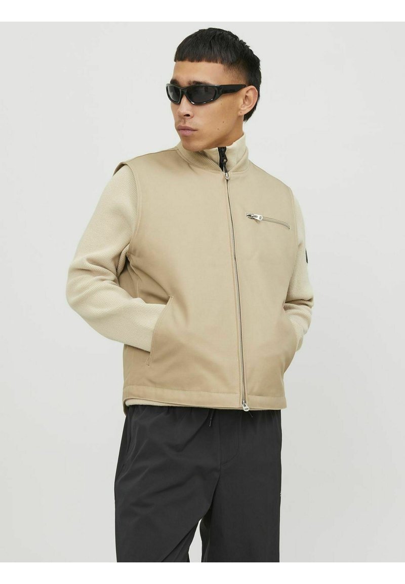 Beige vest met hoge kraag, voorrits en zijzakken. Gladde stof aan de voorkant, gestructureerde mouwen. Model draagt zwarte zonnebril.
