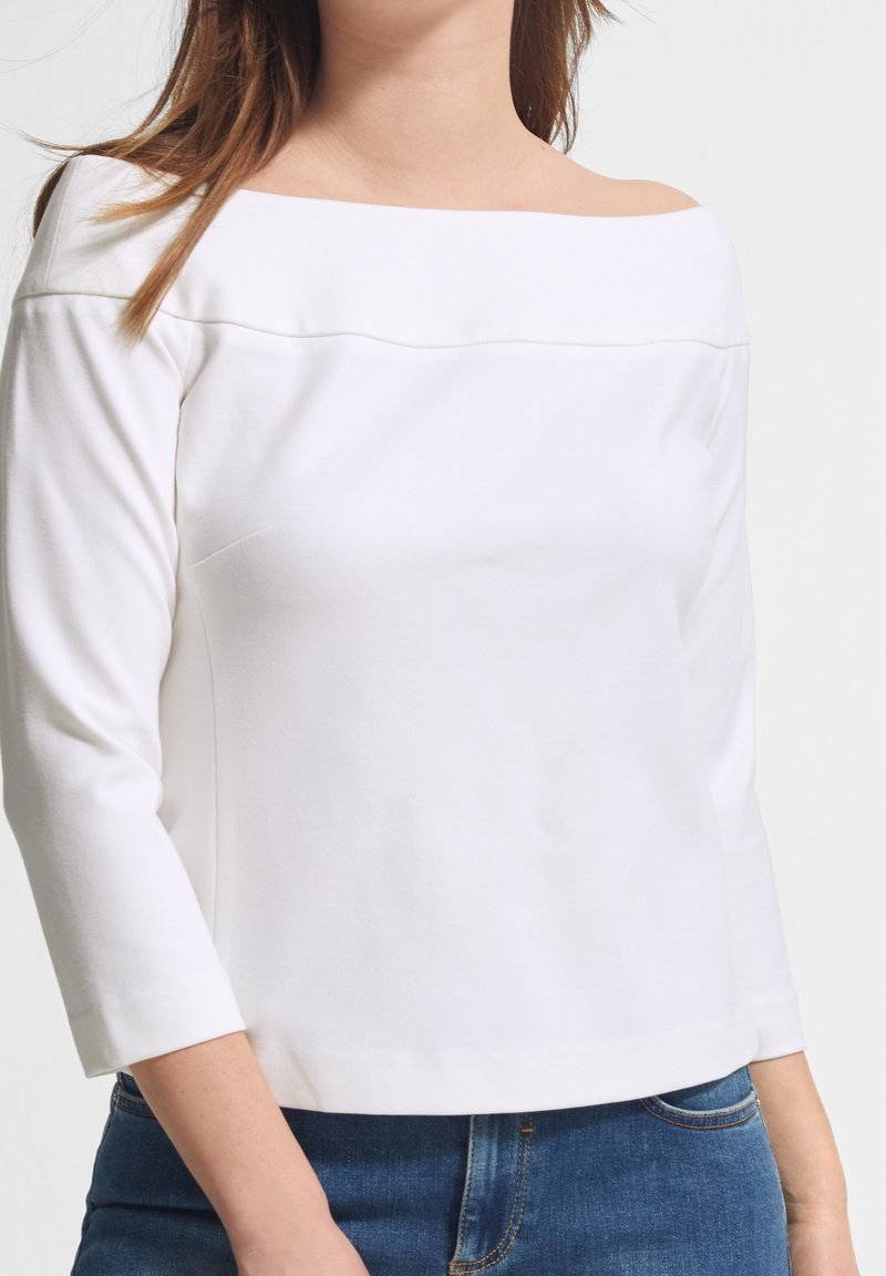Weißes Off-Shoulder-Oberteil aus weichem Stoff, mit Dreiviertelärmeln und geradem Saum, kombiniert mit blauen Jeans.