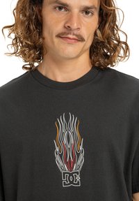 Čierne bavlnené tričko s farebným dizajnom plameňa nad bielym logom. Klasický okrúhly výstrih a krátke rukávy, s hladkou textúrou.