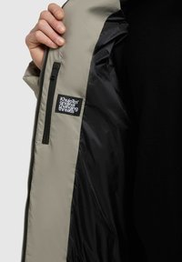Mano che tiene aperta una giacca beige mostrando la tasca interna nera con cerniera e l'etichetta nera con la scritta bianca "Khujo original changing threads".