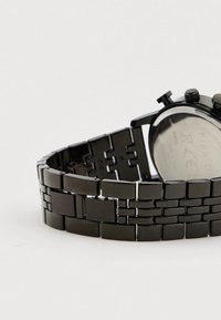 Povezani crni metalni remen sata djelomično zatvoren s vidljivom stražnjom stranom okruglog kućišta sata i dvije bočne tipke.