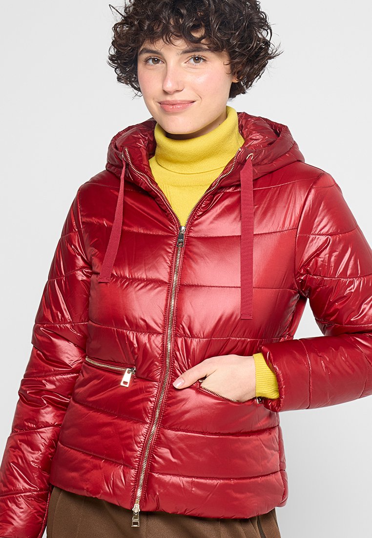 Rode gewatteerde pufferjack met een glanzende afwerking, ritssluiting aan de voorkant, verstelbare capuchon en horizontale stiksels. Draagt over een gele coltrui.