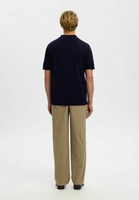 Homme aux cheveux blonds courts, vu de dos, portant un polo bleu marine à manches courtes, un pantalon beige et des chaussures noires sur un fond blanc.