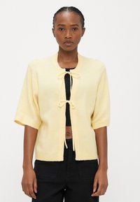 Short-sleeve, yellow knit cardigan with a round neckline, tie closure, and ribbed edges. Paired with a black cropped top and dark pants.