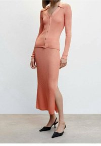 Ensemble en deux pièces en tricot côtelé pêche, composé d'un cardigan à col avec boutons et d'une longue jupe assortie avec une fente sur le côté. Chaussures à talons aiguilles noires avec bride arrière.