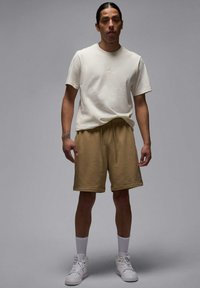 T-shirt in cotone crema con branding, abbinata a pantaloni corti in pile color beige e sneakers bianche. Il modello è in piedi contro uno sfondo neutro.