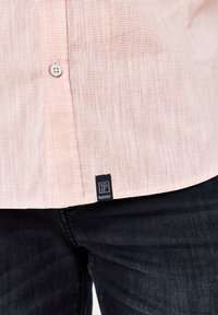 Helles rosa Kurzarmhemd mit einem strukturierten, gestreiften Muster, das einen Knopfverschluss und ein dunkelblaues Etikett mit weißem Text am Saum hat.