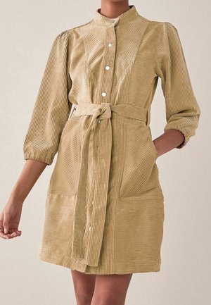 Mujer que lleva un vestido beige de pana con mangas largas, botones a presión en la parte delantera y un cinturón atado en la cintura.