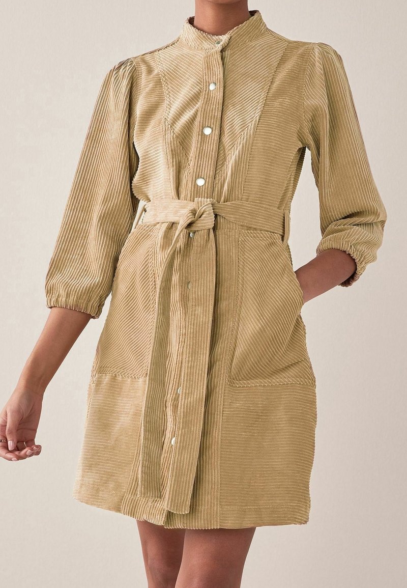 Femme portant une robe en velours côtelé beige à manches longues, avec des boutons-pression à l'avant et une ceinture nouée à la taille.