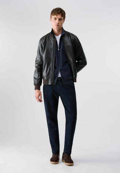 Jeune homme debout portant une veste en cuir noire, un pull zippé bleu marine, un t-shirt blanc, un pantalon foncé et des baskets marron sur un fond clair uni.