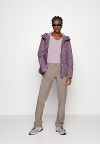 Lichte paarse jas met patroon, lichtpaarse top, beige broek en grijze schoenen. Voorzien van ritsen en een capuchon. Glad textiel overal.