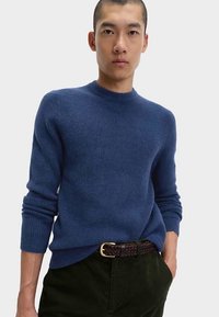 Pull bleu tricoté avec un col rond et une texture côtelée, associé à un pantalon en velours côtelé vert foncé et une ceinture tressée marron.