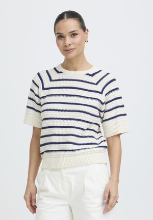 Femeie cu păr închis la culoare, purtând un top tricotat cu mâneci scurte și dungi, și pantaloni albi, stând pe fundal simplu și luminos.