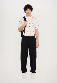 Cream t-shirt with small black heart patterns, paired with black sweatpants, and white sneakers. Model carries a black backpack.