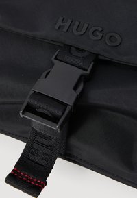 Sac en tissu noir arborant un logo "HUGO" noir bien en vue, une boucle en plastique robuste et une bandoulière texturée avec des accents de couture rouge.
