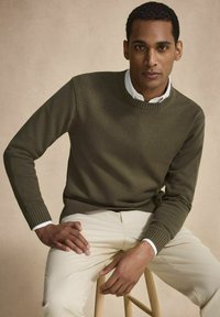 Olivgrön stickad tröja med ribbade ärmslut och nederkant, i kombination med krämfärgade byxor. Modellen sitter på en pall mot en neutral bakgrund.