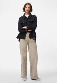 Mlada žena s kratkom kosom u crnoj kordura jakni na kopčanje, bež širokim hlačama i bijelim tenisicama, stoji sa prekriženim rukama.