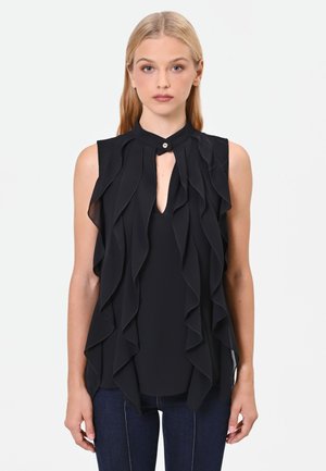 Donna bionda che indossa una camicetta nera senza maniche con strati arricciati e un piccolo bottone al collo alto, abbinata a jeans blu scuro.