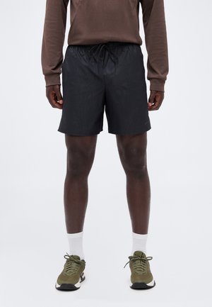 Homme portant un short de sport noir, une chemise marron à manches longues, des chaussettes blanches et des chaussures de course vert olive, debout sur un fond uni.
