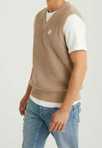 Beige knitted sweater vest with a V-neck, textured fabric, ribbed hem, and a small logo patch on the chest, layered over a white shirt.