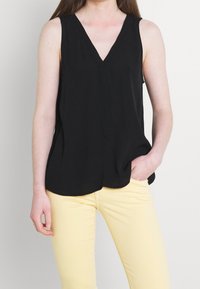 Žena v bezramenném černém topu s výstřihem do V a světlých žlutých kalhotách, s jednou rukou opřenou v kapse na jednobarevném pozadí.