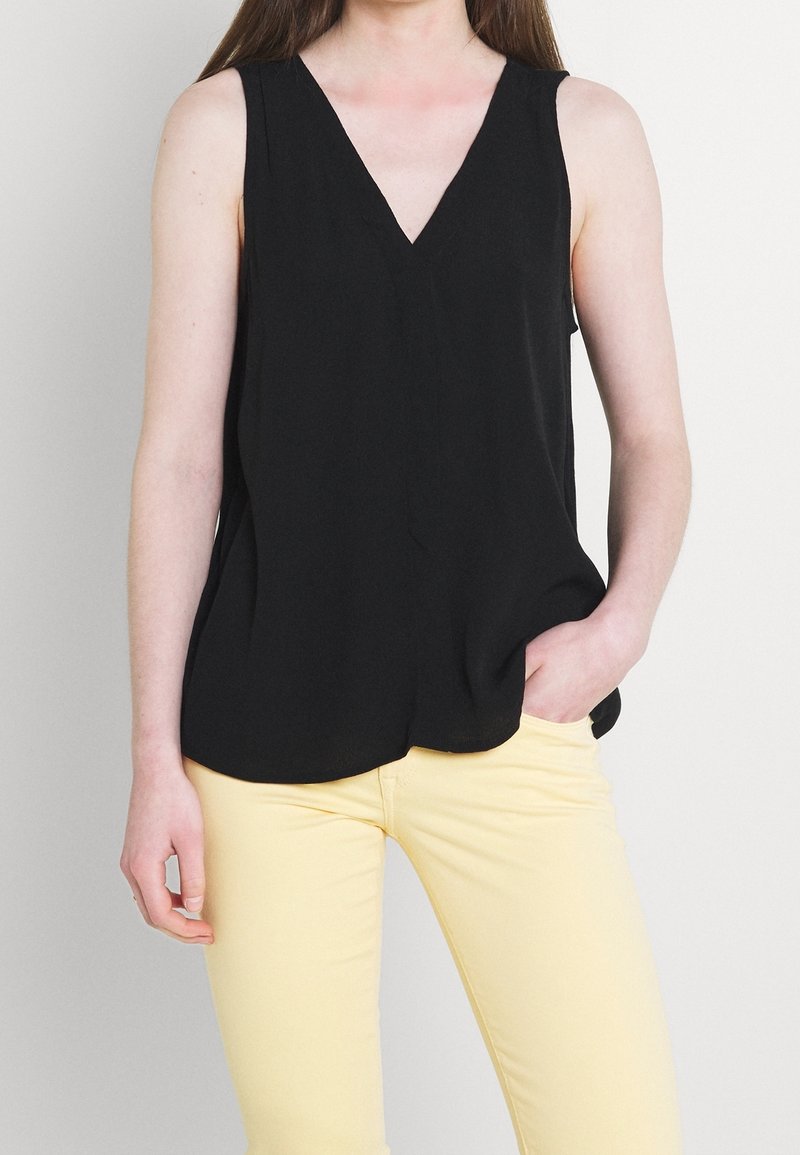 Žena v bezramenném černém topu s výstřihem do V a světlých žlutých kalhotách, s jednou rukou opřenou v kapse na jednobarevném pozadí.