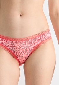 Vrouw in koraalroze, met patronen, kanten ondergoed in bikini-stijl, zichtbare taille en bovenbenen, op een witte achtergrond.
