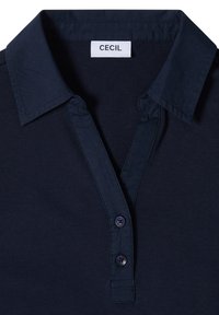 Camiseta tipo polo azul oscuro con dos botones, cuello puntiagudo y una etiqueta blanca que muestra "CECIL" en el cuello.