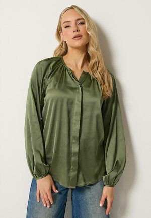Blonde Frau trägt eine olivgrüne Langarmbluse mit gerafften Schultern und blaue Jeans, steht vor einer hellen Wand.